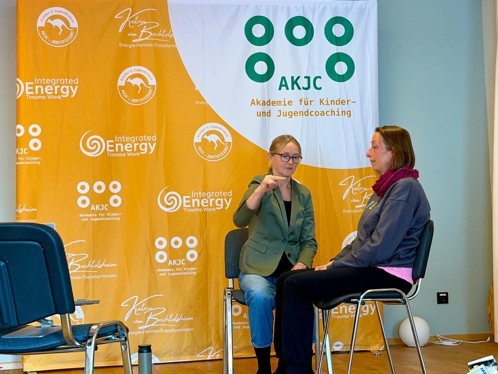 Zwei Personen sitzen sich vor dem AKJC-Banner gegenüber, eine erklärt mit erhobener Hand