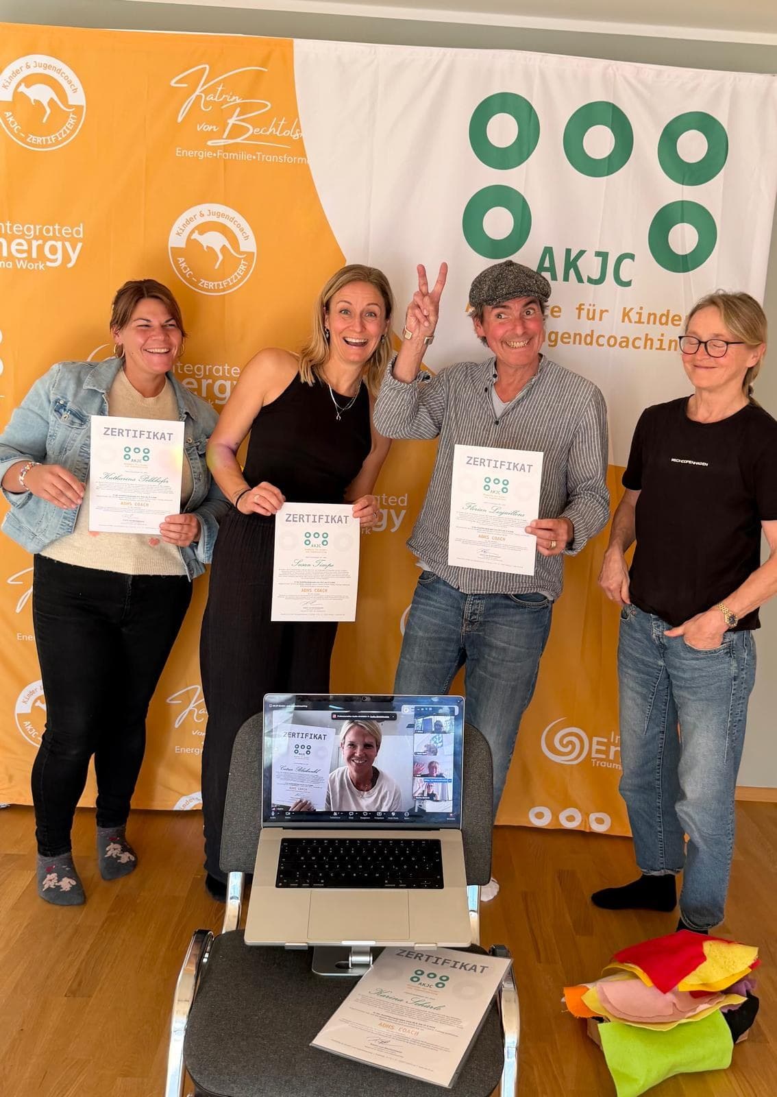 Vier Personen vor dem AKJC-Banner, drei halten ihr Zertifikat «ADHS Coach», vorn ein Laptop mit einer Online-Teilnehmerin, die ihr Zertifikat in die Kamera hält